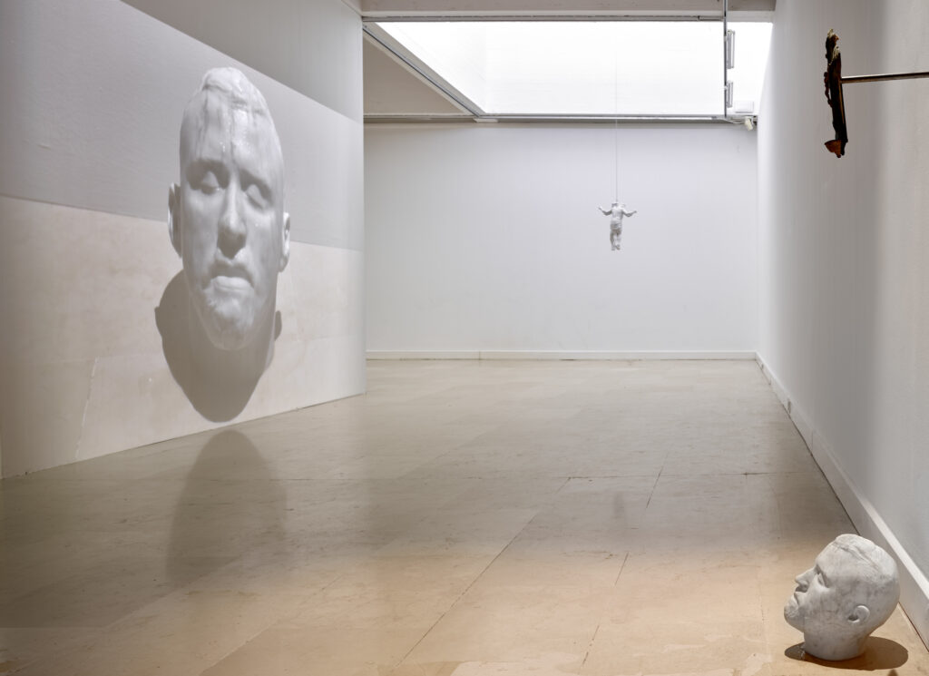 une goutte tombe une sculpture reproduisant le visage de l'artiste, scène filmée et diffusée en direct sur le mur opposé