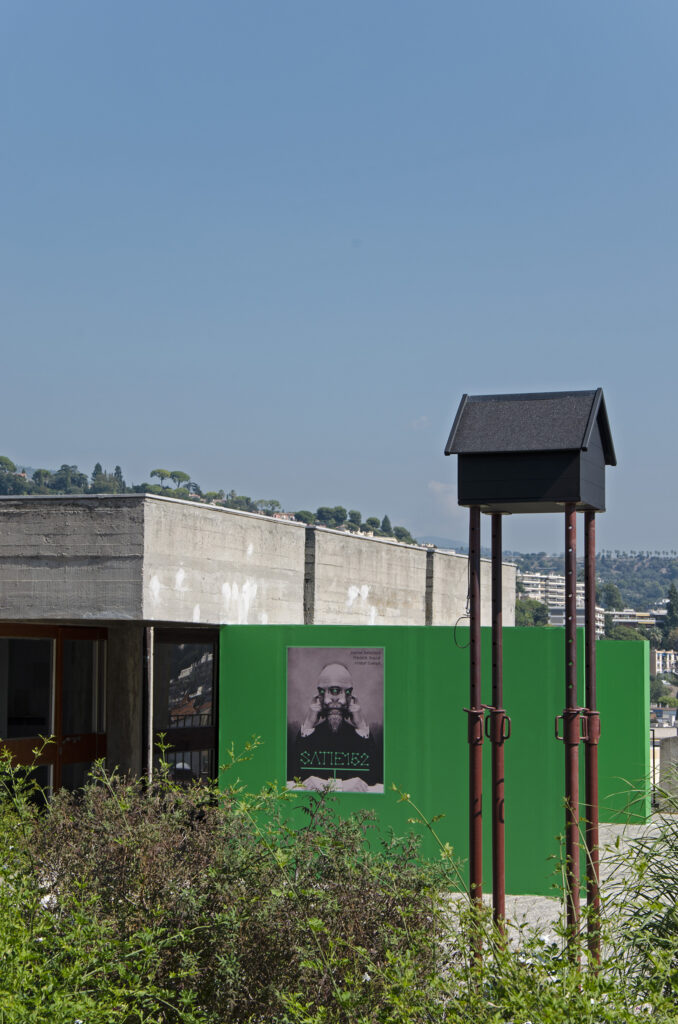 vue extérieure de l'exposition avec une photo de Satie dédoublée sur un mer et un immense perchoir à oiseau en forme de maison