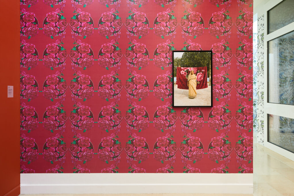vue d'un mur tapissé d'un motif de couverture fleurie en tons de rose sur lequel la photo d'une dame âgée posant devant une couverture au même motif, est accrohéee